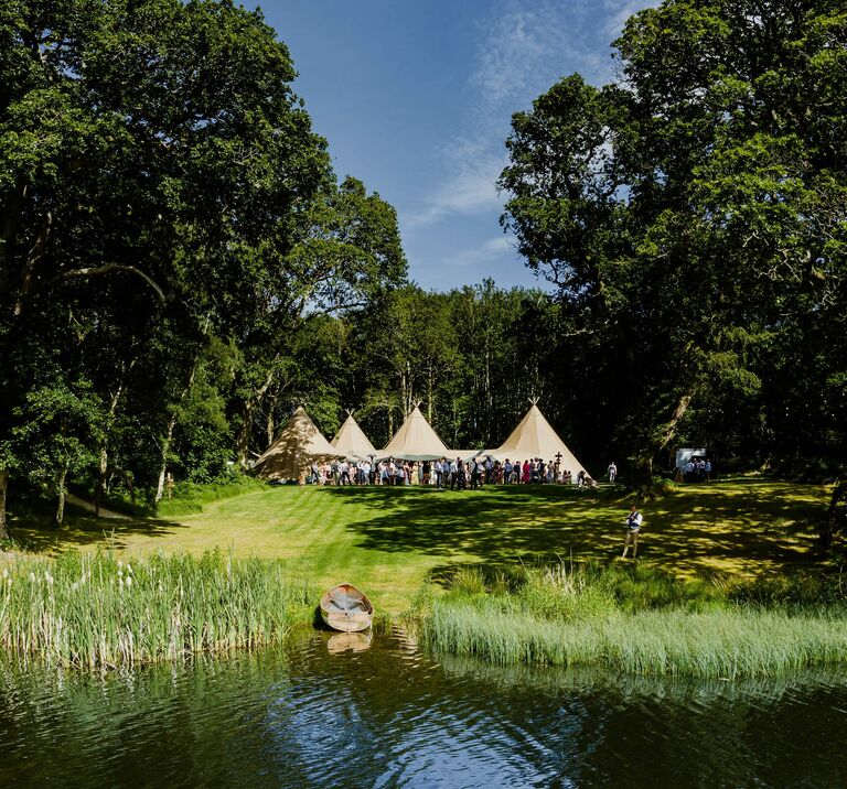 Four tipis sit amongst ancient wood land with a magical lake in the forefront, wedding guests are standing outside celebrating