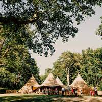 Four tipis sit amongst ancient woodland with a magical lake in the forefront, wedding guests are standing outside celebrating