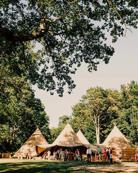 Four tipis sit amongst ancient woodland with a magical lake in the forefront, wedding guests are standing outside celebrating