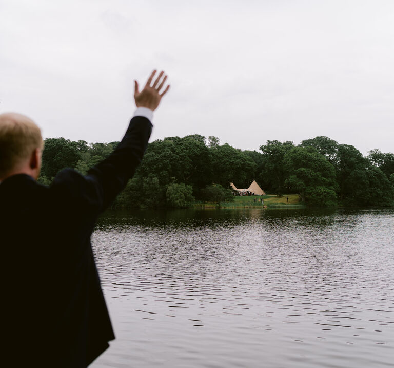 Bride & groom wave to their guests standing at the tipi wedding venue from across the lake