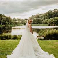 A bride stands with her wedding dress flowing out behind her in front of the lake at the tipi venue at Finnebrogue Woods