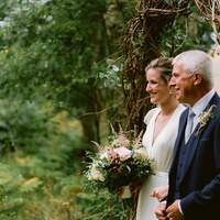 Father and bride walk underneath a willow arch at the start of an aisle