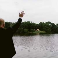 Bride & groom wave to their guests standing at the tipi wedding venue from across the lake
