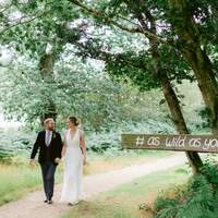Bride & groom hold hands and walk down a path, a wooden signpost saying #aswildasyourlove is nailed to a post
