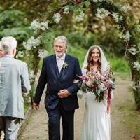 Father & bride walk down the aisle, a wildflower arch stands behind them