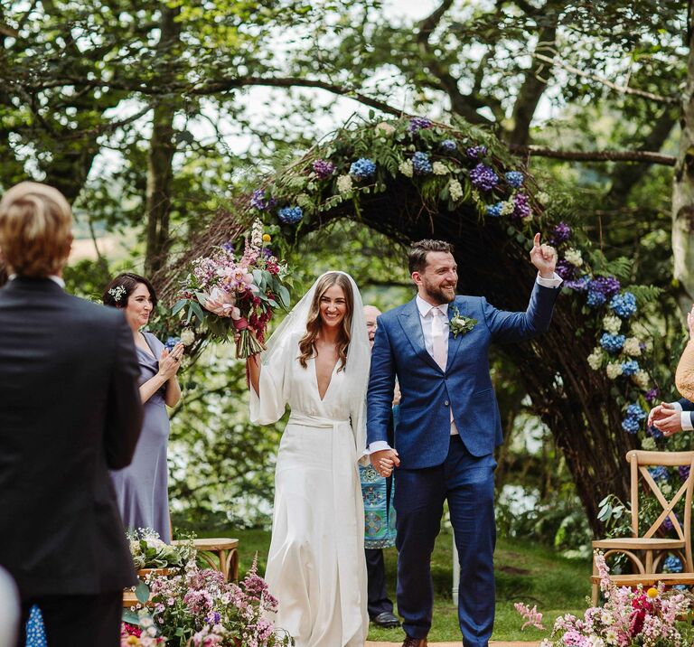 Bride & groom hold hands as they smile & walk down the aisle, the aisle & wicker arch are decorated with wild flowers