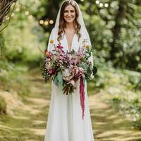 A bride stands below a wicker arch holding a bouquet of wildflowers at Finnebrogue Woods