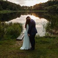 Bride & groom stand together in front of Finnebrogue Woods Lake at sunset