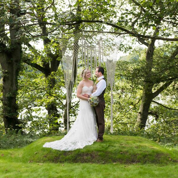 Bride & groom stand under a macrame piece hanging under a tree at our outdoor ceremony site