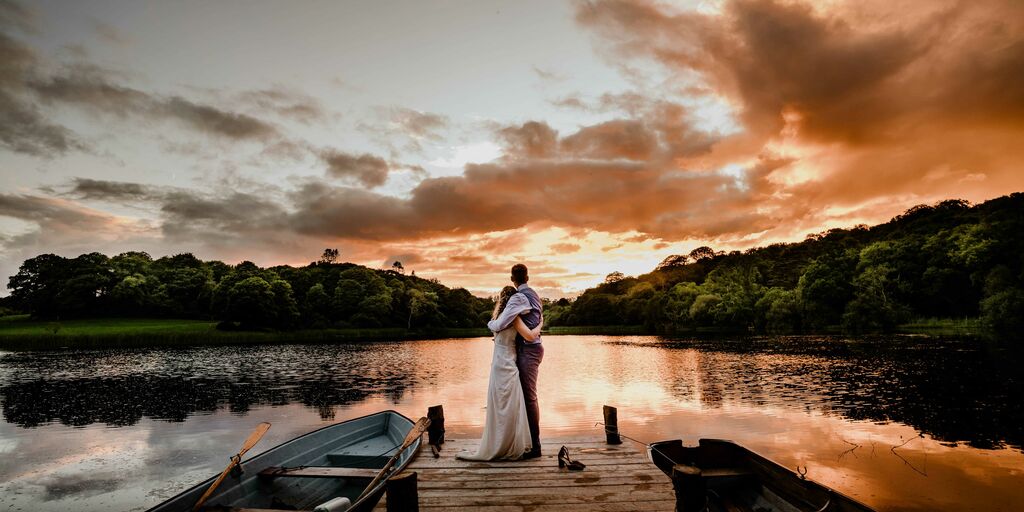 Bride & groom stand on a jeti overlooking Finnebrogue Woods and the Lake at sunset, two rowboats sit either side of the jeti