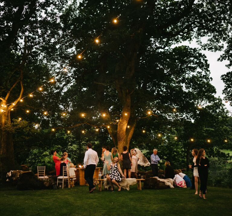 Wedding guests gather around the firepit area in the trees at Finnebrogue Woods, festoon lighting hangs above them