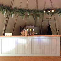 A wooden bar stands inside the tipi wedding venue, crossbeams are dressed in wild greenery