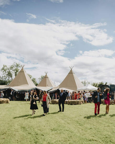 Guests stand outside talking with a four tipi Wedding reception on an open field, square hay-bales sit in front