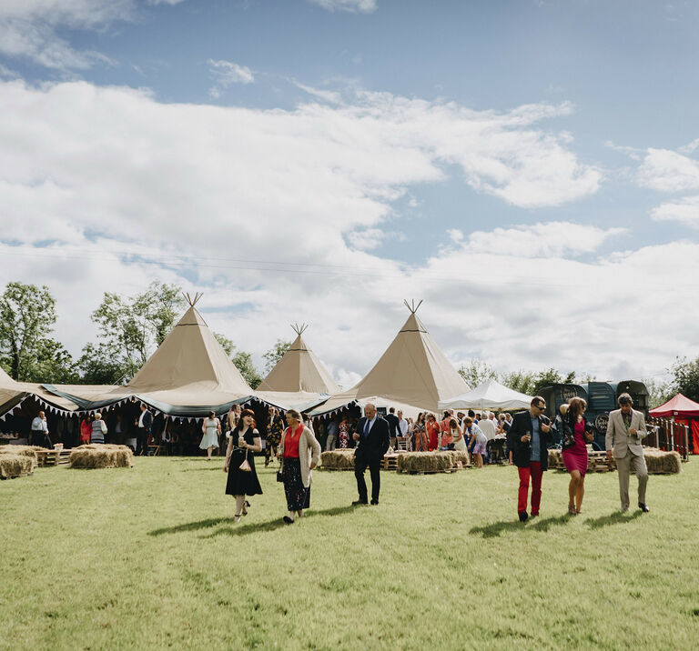 Guests stand outside talking with a four tipi Wedding reception on an open field, square hay-bales sit in front
