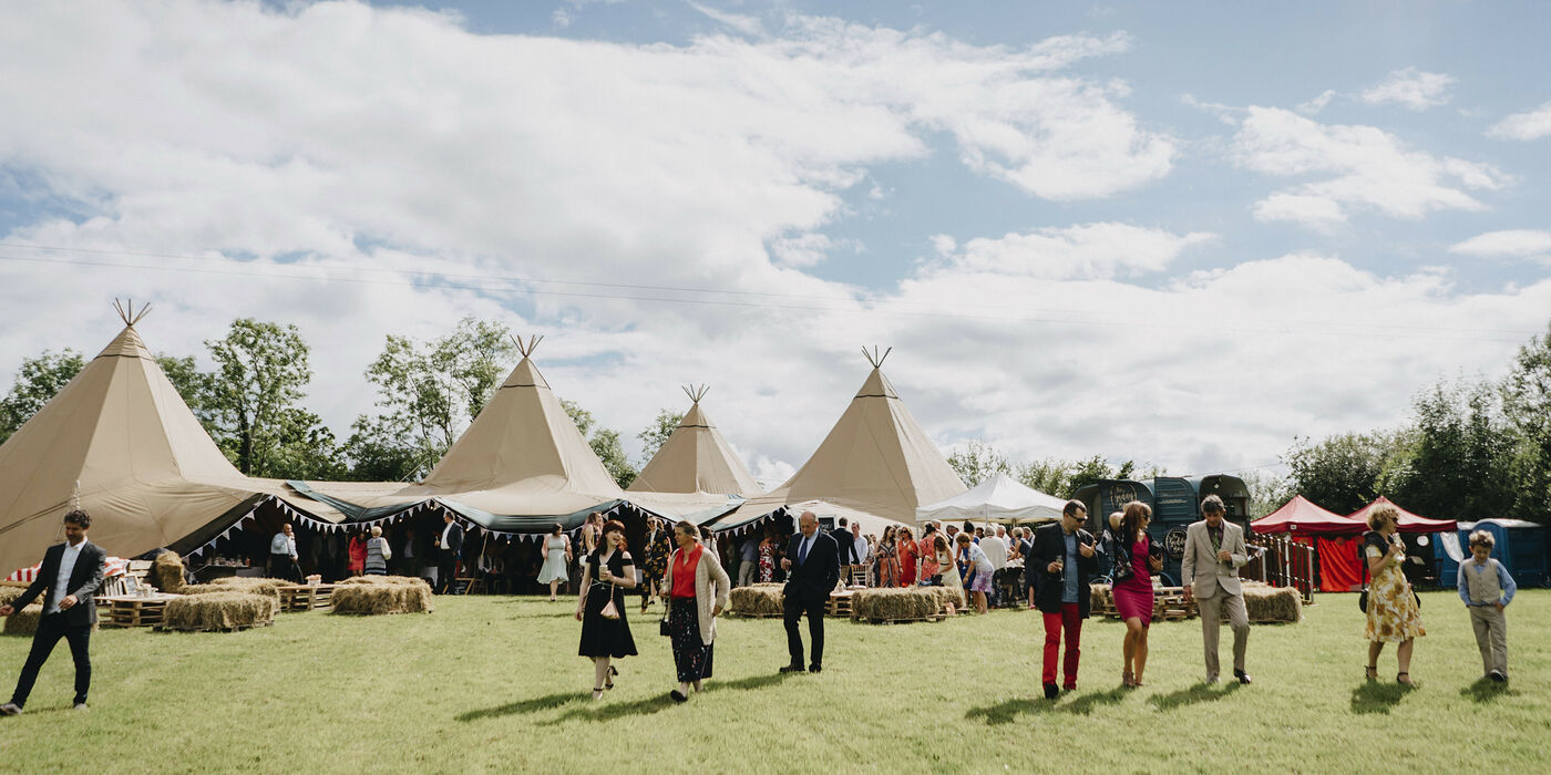 Guests stand outside talking with a four tipi Wedding reception on an open field, square hay-bales sit in front