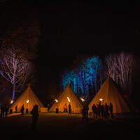 Three Tipis and the trees behind them are lit up with uplighters, corporate guests are silhouetted by fire torches