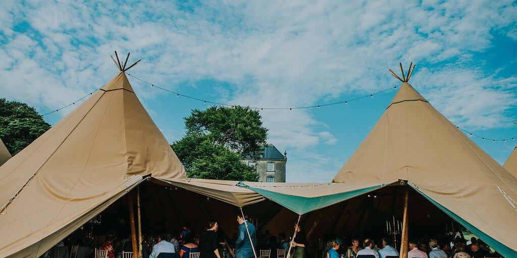 Open sided entrance to a tipi wedding reception, inside is crowded with guests sitting at tables