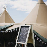 A blackboard easel welcomes guests to the wedding, Tipis sit behind it with bunting decorating the entrance